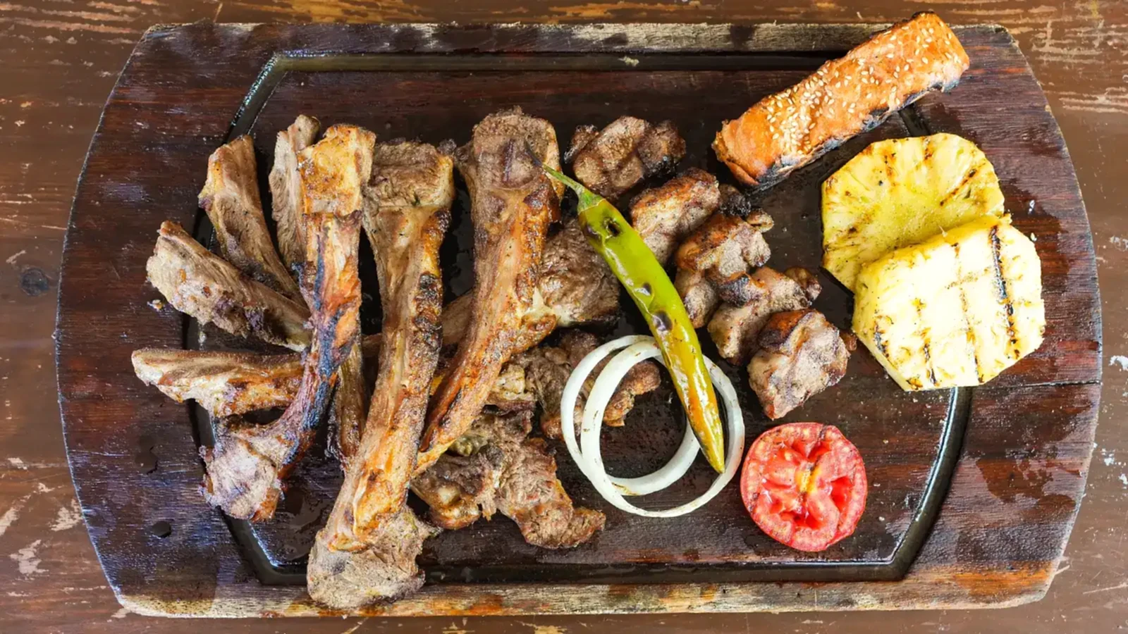 Kömür ateşinde pişirilmiş kuzu kafes ızgara, yanında közlenmiş biber, domates, halka soğan ve ananas dilimiyle döküm tabakta sunum – Bakı Dede Kebap Steak Burger Tatlı Kasap